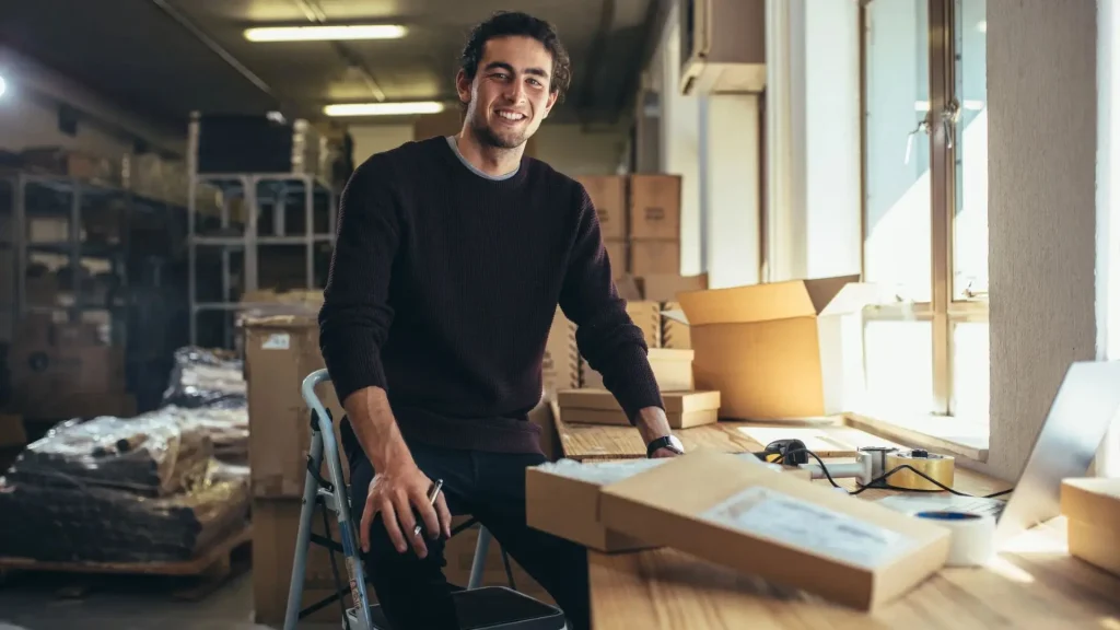 Smiling man in a dark sweater sits at a cluttered desk in a warehouse with boxes and packing materials around him.